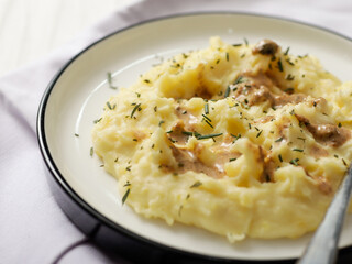 A plate of creamy mashed potatoes topped with a rich, savory gravy and garnished with fresh herbs. The dish is served on a white plate, with a spoon placed on the side, ready to be enjoyed