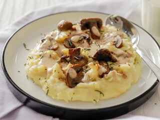 A plate of creamy mashed potatoes topped with a rich mushroom gravy and garnished with fresh rosemary. The dish is elegantly presented on a white plate, highlighting the hearty, comforting meal.