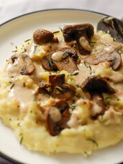 A plate of creamy mashed potatoes topped with a rich mushroom gravy and garnished with fresh rosemary. The dish is elegantly presented on a white plate, highlighting the hearty, comforting meal.
