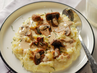 A plate of creamy mashed potatoes topped with a rich mushroom gravy and garnished with fresh rosemary. The dish is elegantly presented on a white plate, highlighting the hearty, comforting meal.