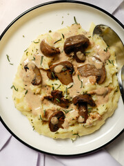 A plate of creamy mashed potatoes topped with a rich mushroom gravy and garnished with fresh rosemary. The dish is elegantly presented on a white plate, highlighting the hearty, comforting meal.