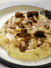 A plate of creamy mashed potatoes topped with a rich mushroom gravy and garnished with fresh rosemary. The dish is elegantly presented on a white plate, highlighting the hearty, comforting meal.