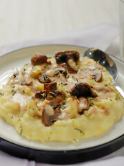 A plate of creamy mashed potatoes topped with a rich mushroom gravy and garnished with fresh rosemary. The dish is elegantly presented on a white plate, highlighting the hearty, comforting meal.