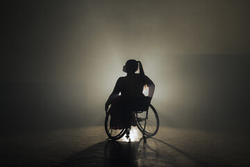 Obraz premium Graceful wheelchair dance of a woman with a disability on a black background with bright backlight. Para dance concept.