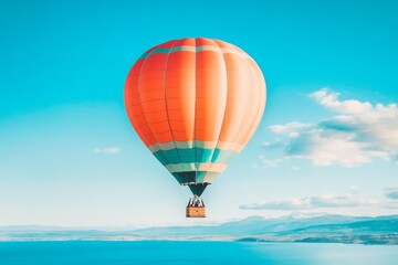 Fototapeta premium Vibrant orange and blue hot air balloon gracefully floating in a serene, clear blue sky over a vast, tranquil body of water with fluffy white clouds in the far distance.