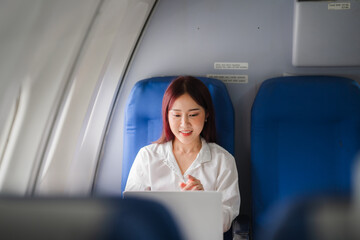 Productive Journey: A young Asian businesswoman smiles gently as she works on her laptop in the bright cabin of an airplane, showcasing the ease of modern business travel. 