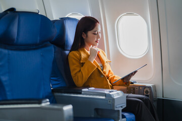 Businesswoman on Airplane: A young businesswoman in a yellow blazer sits by the window of an airplane, engrossed in her tablet. She is on a business trip, and her expression is one of focus and determ