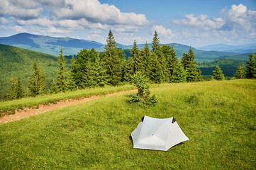 White tourist tent on sunlit, grassy hilltop, surrounded by young pine trees. Panoramic view of lush, rolling mountains under bright blue sky with scattered clouds, creating idyllic camping spot.