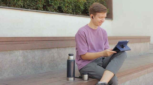 Young man sitting on bench and watching video with headphones on tablet.
Programmer on lunch break in the park.