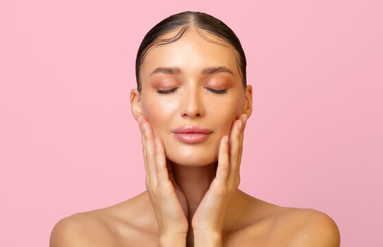 Beautiful lady with perfect skin touching her cheeks with pleased face expression, enjoy result of treatment over pink background