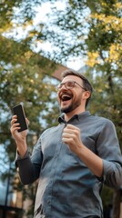 A happy man in casual looking at his phone and celebrating a victory, with an office building in the background