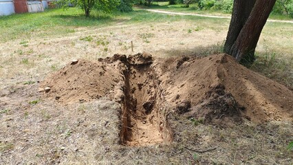 A trench has been dug along tree trunks in a grassy lawn to lay pipes, cables and other utilities. Next to the trench is excavated soil. A building stands nearby. Sunny summer weather and shadows