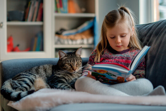 little girl reading a book next to her cat on the couch