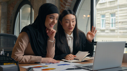 Two multiracial businesswomen Asian Korean woman girl Muslim Islamic hijab female businesswoman in office work video call laptop virtual conference paperwork diagram result present business women