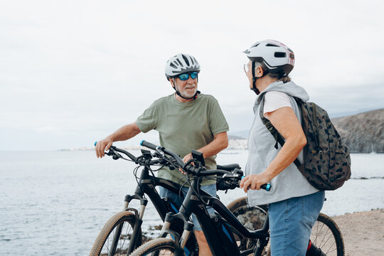 Senior couple riding bikes together in rocky beach enjoying outdoor. Active mature people talking holding mountain bikes. Bicycle lifestyle concept, having fun.