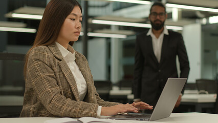 Asian woman worker female girl businesswoman intern at office working laptop computer pc typing online Indian business man guy male leader boss businessman go help partnership multiracial colleagues