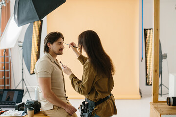 Stylist woman applying make up to handsome model at photo shoot backstage, getting ready for...