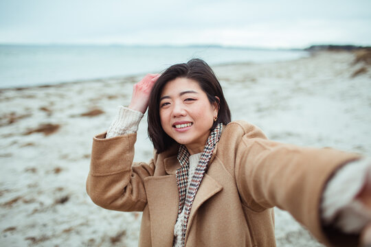 Smiling young Asian woman taking selfie on beach during winter