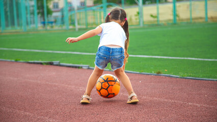 Little player: child girl playing with soccer ball at soccer field in summer day outdoors. Child...