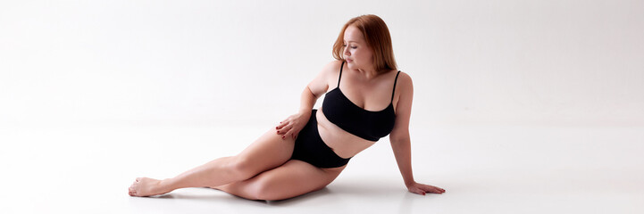 Fototapeta premium Calm and relaxed young beautiful woman with overweight body shape posing on floor in underwear isolated on white studio background. Concept of female beauty, body positivity, care, acceptance