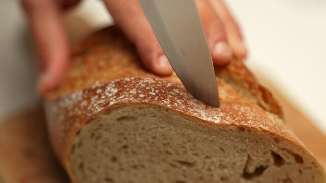 On a wooden breadboard, fresh, appetizing bread is placed alongside a sharp knife. A man&rsquo;s hands enter the frame, slicing the bread with the knife.