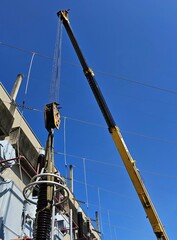 A close-up of a worker using a crane to install a large, ceramic electrical insulator on a high-voltage power transformer. The insulator is suspended by a hook attached to the crane's cable. The trans