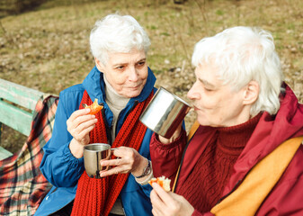 Two elderly women are sitting on a bench in the park, drinking coffee, eating pizza, relaxing and enjoying life.