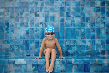 A young boy sitting on the edge of a swimming pool, wearing a blue swim cap and goggles, ready to swim