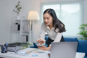 Young asian businesswoman calculating and taking notes on her smartphone while working on a project...