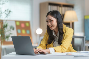 Young asian businesswoman is working on a laptop in her modern office, smiling at the screen