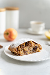 Freshly baked powdered apple strudel with raisins on white plate with red apples and cinnamon on white background. homemade Apfelstrudel, austrian germany cuisine. Menu, recipe. Close up, vertical