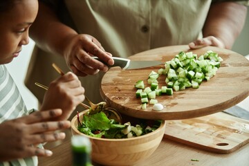 Close up on hands of Black senior woman adding chopped cucumbers to green lettuce leaves in salad bowl using wooden cutting board and knife while cooking together with girl at kitchen, copy space
