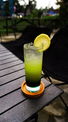 A refreshing green drink with a lemon wedge garnish sits on a wooden coaster on a table outdoors.