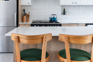 Kitchen island with wood look cabinets, white stone counter top surface and wood and green bar chairs, blurry background