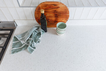 Styled surface on a modern white stone kitchen counter with white vertical wall tiles. Wooden cutting board, green kitchen towel cloth, small bowls and bottle of olive oil