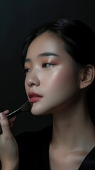 Fototapeta premium Asian woman applies blush with kabuki brush in beauty studio. Elegant updo and dark background highlight makeup process. Perfect for beauty promo or editorial use.