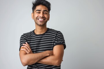 Fototapeta premium Young Indian man wearing black striped t-shirt stands confidently on isolated white background. Happy face smiling with crossed arms looking directly at camera. Positive, cheerful person expressing