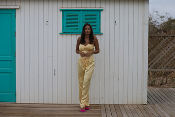 Young and beautiful brunette woman poses self-confident in front of the white and turquoise wooden hut on a beach in Cadiz, andalusia, Spain. The woman makes different body postures like a model.