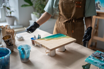 Unrecognizable female artist applies paint on epoxy with long stick, designing pattern for future artwork on wooden board in workshop. Creative occupation, art idea, small business, do it yourself