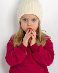 Portrait of beautiful cute little toddler girl. Child with funny face in burgundy color sweatshirt and white hat. Pretty smile kid in studio on light background. Vertical photo