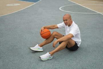 Fototapeta premium Basketball player or sporty man sitting, looking at camera and rest with ball on sports court at courtyard. Urban style. Copy space.