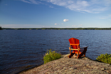 An empty red Adirondack chair on a rocky shoreline along the lake, Muskoka Lake, Ontario, Canada 