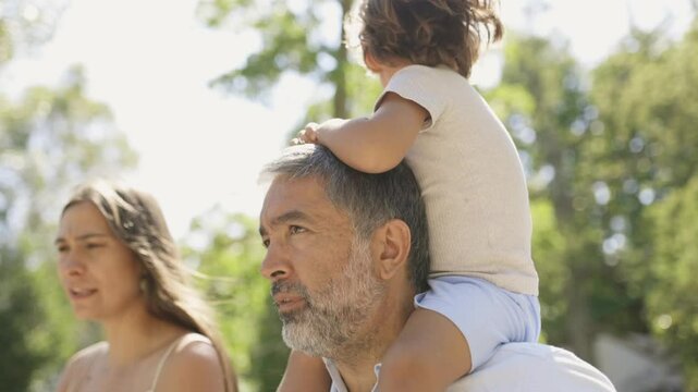 Grandfather is carrying his grandson on his shoulders while walking in the park with his family