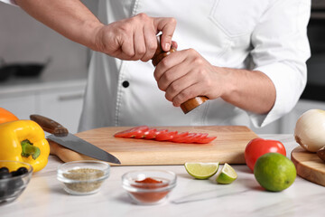 Professional chef adding salt to tomato at white marble table in kitchen, closeup