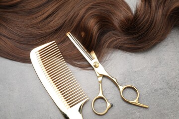 Brown hair strand, comb and professional scissors on grey surface, flat lay