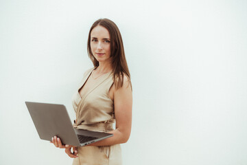 Naklejka premium Portrait of a confident young hispanic woman holding a laptop against a plain light-colored background. She is wearing a beige dress, standing slightly angled, looking directly at the camera