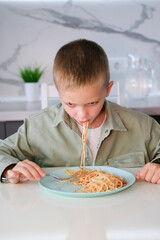 Boy eats spaghetti with appetite. Funny child gets dirty eating pasta, wraps it on fork from large...