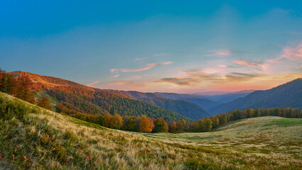 A serene autumn landscape with a cluster of vibrant trees on a hillside overlooking a vast range of blue-tinged mountains. The sky transitions from soft hues to the golden light of dusk