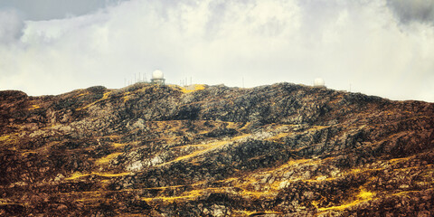 The image shows a rocky, barren landscape with visible signs of erosion. The peak is capped by two...