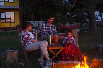 Naklejka premium Family is sitting by the outdoor campfire in the courtyard of the house on camping chairs, drink tea from a thermos, warm up and chat by the hearth